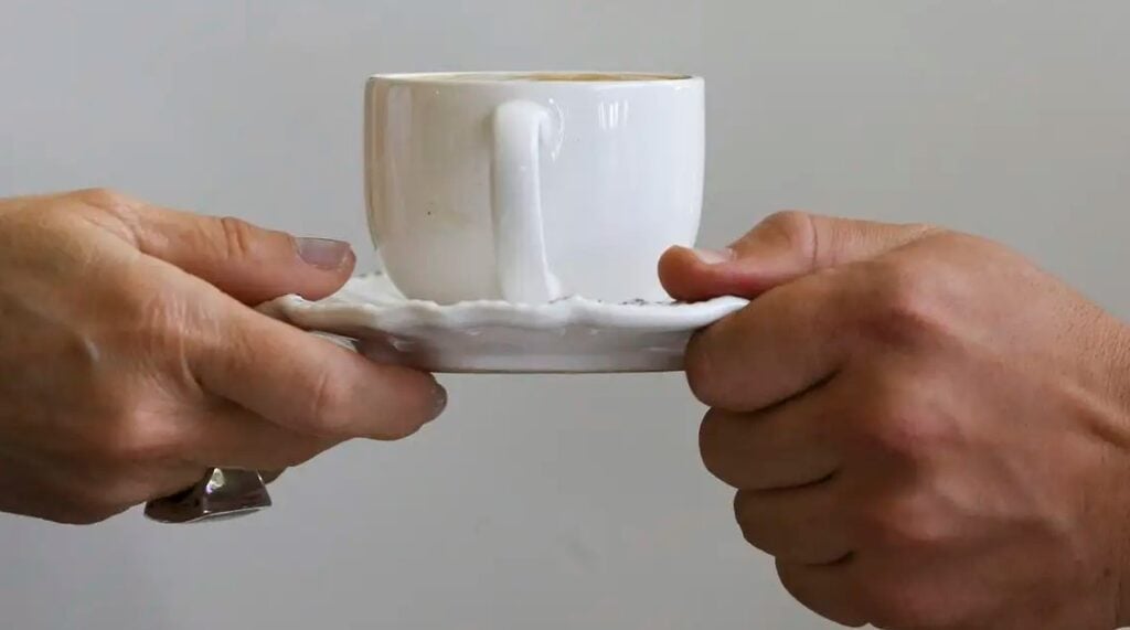 Imagem mostra duas mãos segurando uma xícara de café e o pires branco